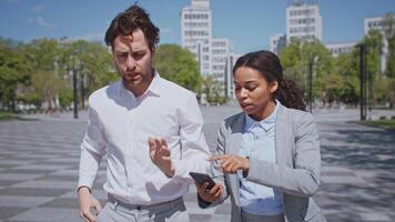 A man and woman, both young professionals, converse outdoors about business. They are dressed in formal attire and focus on their smartphones while standing in a modern urban environment. video
