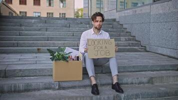 A young Caucasian man sits on steps holding a sign that says he is looking for a job. Beside him is a box filled with office supplies and plants, reflecting a search for new career paths. video