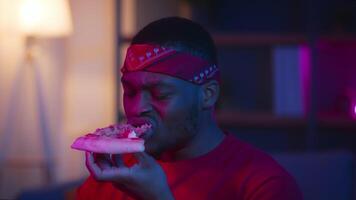 A millennial man with a bandana takes a break from his remote work to enjoy pizza. Neon lights create a futuristic atmosphere in his home office as he indulges in a late-night snack. video