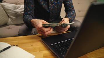 Man counting dollar banknotes, sitting in front of coffee table with laptop and notebook. Person managing budget at home, counts money in living room video