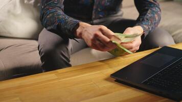 Man holding one hundred euro banknotes, sitting on sofa in front of table with open laptop. Person counting cash money at home. Personal finance and home budget concept video