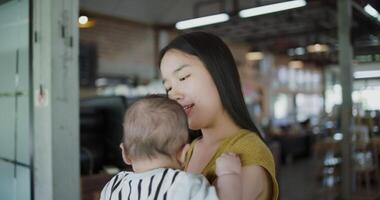 Handheld shot, Young asian mother stands in a coffee shop, smiling warmly as she gently cradles a baby boy in her arms with a relaxing atmosphere during happy time video