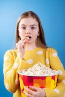 Girl eating popcorn from a colorful bucket, wearing a yellow sweater, enjoying snacks with a surprised expression on a blue background photo