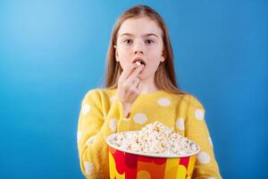 Girl eating popcorn from a bucket, wearing a yellow sweater, enjoying snacks with a surprised expression on a blue background. photo