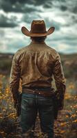 Cowboy standing in a wildflower field under a cloudy sky with a rugged mountain backdrop photo