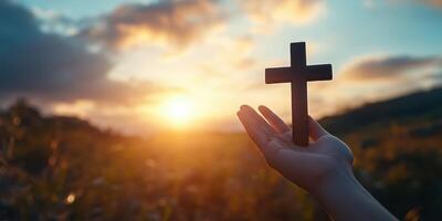 Hand holding a wooden cross against a vibrant sunset in a serene landscape photo