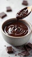 Rich melted chocolate being poured from a wooden spoon into a textured gray bowl on a light surface photo