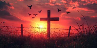 Sunset over a cross silhouette with birds flying in the vibrant sky and barbed wire in a tranquil field photo