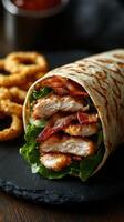 Delicious chicken wrap with crispy bacon and fresh lettuce served on a black plate alongside onion rings photo