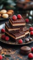 Delicious chocolate dessert squares topped with fresh raspberries on a dark plate surrounded by sweets photo