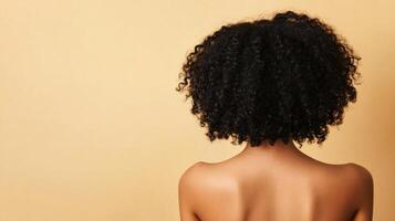 A woman with dark curly hair faces away from the camera showing off her natural texture and a smooth dark complexion against a light tan background. photo