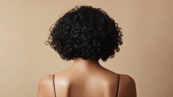 Curly-haired woman. The dark curls add volume. She is seen from the back, wearing a minimalist top, against a soft beige backdrop. photo