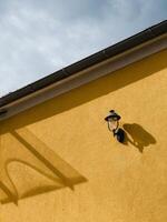 Outdoor lamp casts a shadow on a vibrant yellow wall, complemented by a gutter and a cloudy sky, creating a striking contrast of light and shadow in an urban setting. Lamp and bright yellow wall photo
