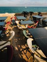 Seaside restaurant terrace featuring empty tables, vibrant chairs, sailboat gliding across blue horizon with calm ocean backdrop photo
