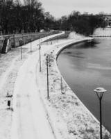 Snow covered promenade lining the river in winter, featuring street lamps, benches, and trash cans, captured in striking black and white image that evokes tranquility and solitude. promenade in winter photo