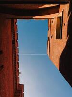 Blue sky between red brick defensive walls. Low angle view of airplane contrail crossing blue sky between high red brick defensive walls with merlons, creating a sense of enclosure and strength photo