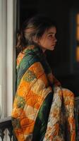 Young woman wrapped in colorful quilt gazing out the window in a cozy room during early evening hours photo