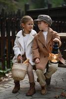 young boy and girl sit closely on a rustic bench dressed in timeless vintage attire. the warm glow of their lantern contrasts and basket beside them adds charm of their nostalgic moment photo
