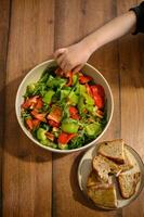 A bowl of fresh vegetable salad with croutons sits on the table as small hands reach for it. photo