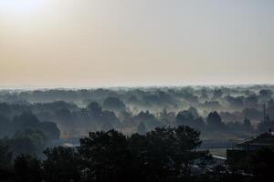 foggy landscape, above deciduous forest, early in the morning. houses in the distance and the surface of the river are visible between the trees. the sun is reflected in the surface of the water. fog to the horizon photo
