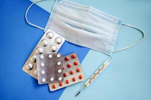 tablets in a blister, a medical mask and a thermometer on a blue background. three blisters of tablets photo