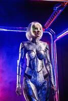 Blonde as a cyber robot. Girl in shining silver armor in neon. She walks in a glowing cube, smoke around. Blonde with fantastic makeup and image in a dark key and colored light, posing in cyberspace photo