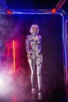 Blonde as a cyber robot. Girl in shining silver armor in neon. She walks in a glowing cube, smoke around. Blonde with fantastic makeup and image in a dark key and colored light, posing in cyberspace. photo