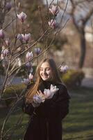 young blonde girl and blooming pink magnolia. happy girl laughing in the sun rays. she is standing near a blooming tree in the park. she has long straight hair and a dark coat. photo