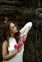 charming girl with roses in her hands in nature against the backdrop of rocks. with fashionable makeup and dark wavy hair posing in nature. in a white bodysuit with a slanted neckline. in the background a waterfall photo