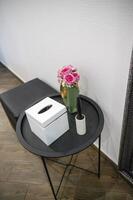 round black metal table next to it pouf. on the table are a box for napkins, decorative flowers, a brush for cleaning clothes. vertical, top view. on the floor are wood-like tiles. the walls are white photo
