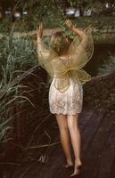 cute blonde girl in fairy costume with wings. girl in nature reeds grow around. fairy wings of golden color, made of transparent chiffon, decorated with embroidery. concept of magic and nature. back view, full length photo
