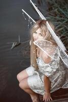 cute blonde girl in fairy costume with wings. girl sitting on pier near river. fairy wings white, made of transparent chiffon, decorated with pearls. concept of magic and nature. top view. looking at camera photo
