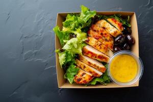 Grab-and-go salad with lettuce, grilled chicken, black olives, and lemon dressing in a cardboard box on a dark textured background. Fresh and healthy takeaway meal. . photo