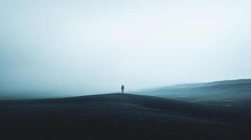 Solitary figure standing atop a dark, desolate hill enveloped in fog, evoking a sense of mystery and solitude in the monochromatic landscape. photo