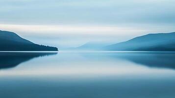 Serene waters mirror misty mountains, creating a calming, monochromatic landscape. The horizon fades into the sky, evoking tranquility. photo