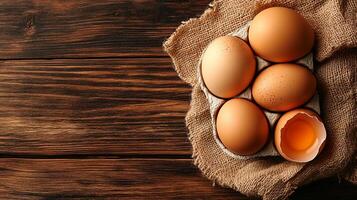 Fresh brown eggs in carton atop burlap on rustic wooden table. One cracked open, yolk visible. Natural light creates soft shadows. photo