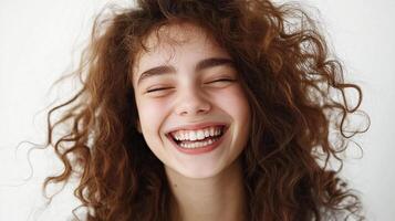 Radiant young woman with curly hair expressing pure joy with closed eyes and a wide, genuine smile against a clean, light background. photo