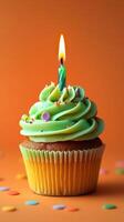 Colorful birthday cupcake with green frosting and a lit candle set against a vibrant orange background photo