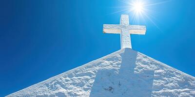 White cross atop a hill under a bright blue sky with sun shining in the background photo