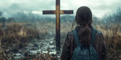 Girl with wet hair stares at a wooden cross in a desolate field during a rainy day at dusk photo