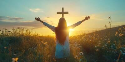 Woman in white dress stands with arms outstretched towards sunset holding a cross in a field of flowers photo