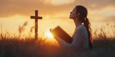 Woman in prayer at sunset near cross while reading a book in an open field photo