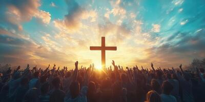 Large crowd raising hands in worship during sunset behind a prominent cross in a serene sky photo