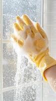 de cerca de un mano en amarillo caucho guante limpieza un ventana con jabón burbujas foto