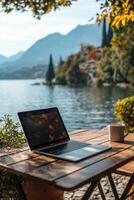 Laptop on a wooden table overlooking a serene lake with mountains in the background photo