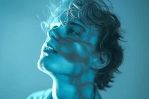 A young Caucasian male with tousled hair gazes upwards, illuminated by soft blue lighting that casts striking shadows on his face. photo