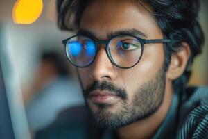 A focused South Asian man with glasses gazes intently at a computer screen, immersed in thought. photo
