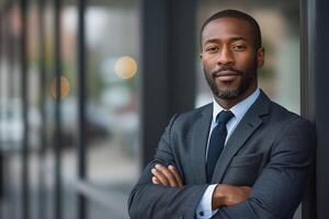 Confident Black man in business attire, standing with crossed arms near glass windows, exuding professionalism and determination. photo