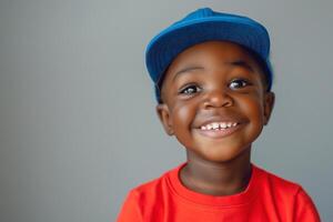 A joyful Black boy, around 5 years old, wearing a red shirt and blue cap, smiles brightly against a gray background. photo