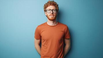 Portrait of a red-haired man with glasses, wearing an orange shirt, standing against a blue wall with a neutral expression on his face. photo